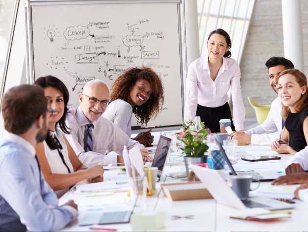 A diverse group of professionals meet in a modern boardroom, collaborating around laptops and documents with a whiteboard full of strategy notes in the background. The scene reflects how in-house counsel and business leaders work together to balance innovation, compliance, and AI governance in corporate decision-making.